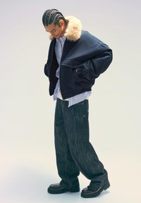 Marinfärgad bombarjacka med en krage av fuskpäls, lagd över en ljusblå skjorta, parat med mörk denimjeans och svarta klumpiga skor.