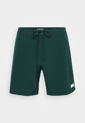 Grønne svømmebukser laget av lett materiale, med snøring i livet, sidelommer og et lite hvitt logo-patch.