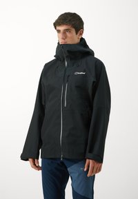 Jachetă neagră, impermeabilă, cu guler înalt și glugă ajustabilă, având un fermoar frontal și un logo pe piept. Purtați-o cu pantaloni albaștri.