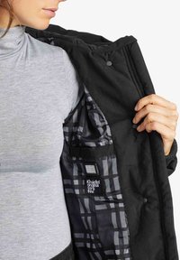 Svart jacka med rutigt foder, med en slät ytteryta. Insidan har en ficka med en etikett. Användaren håller den öppna jackan.