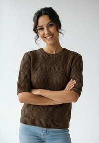 Uśmiechnięta kobieta o ciemnych włosach, ubrana w brązowy, krótki sweter z dzianiny w warkocze i jasnoniebieskie dżinsy, ze skrzyżowanymi ramionami na neutralnym tle.