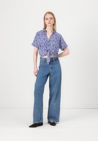 Blå mønstret kortermet skjorte knyttet i livet, kombinert med vide lyseblå jeans og svarte leppestift. Enkle tilbehør fullfører stilen.
