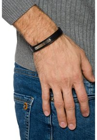 Czarna skórzana bransoletka z gładką metalową płytką i dwoma srebrnymi ćwiekami, noszona na ręce w zestawieniu z szarym swetrem i niebieskimi dżinsami.