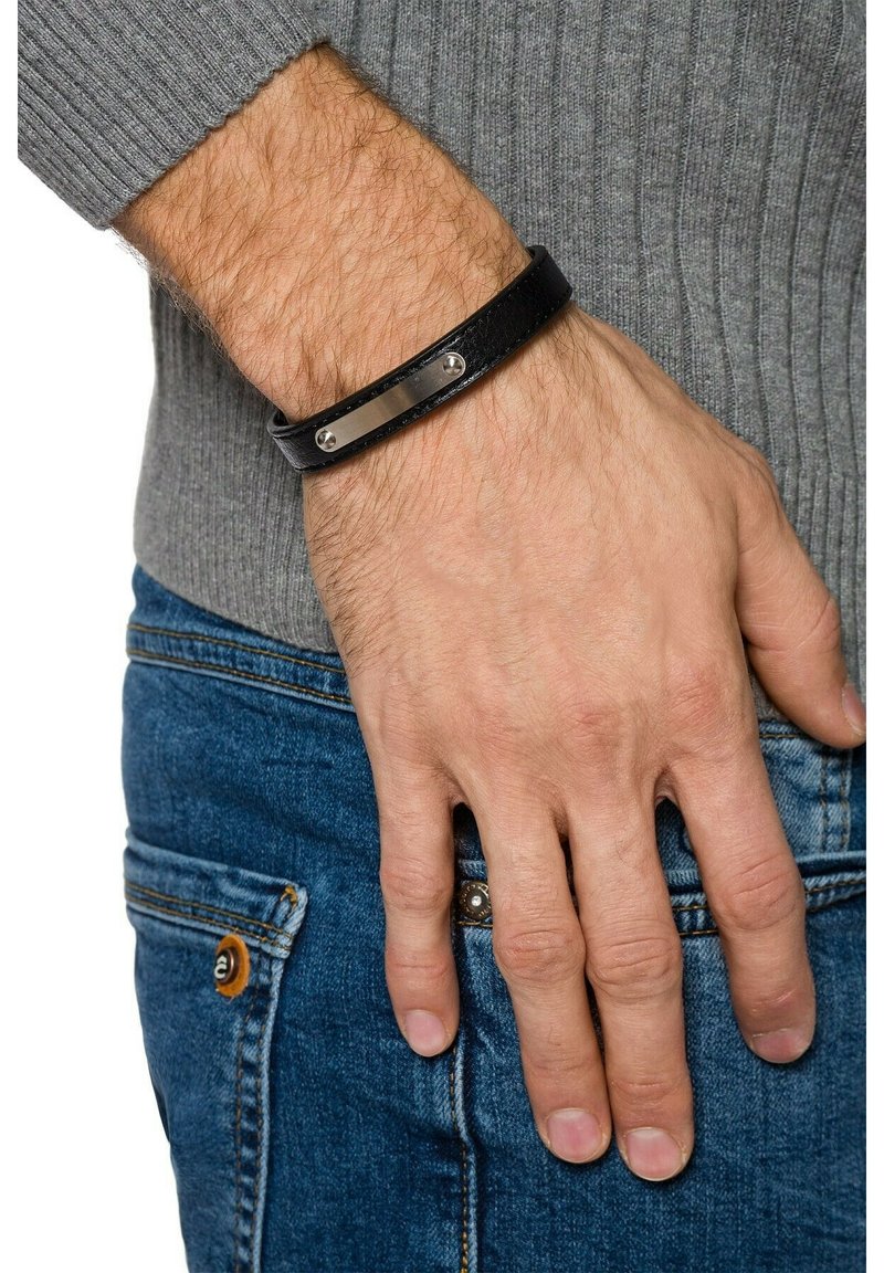 Czarna skórzana bransoletka z gładką metalową płytką i dwoma srebrnymi ćwiekami, noszona na ręce w zestawieniu z szarym swetrem i niebieskimi dżinsami.