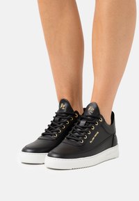 Černé kožené tenisky se zlatými akcenty, bílé gumové podrážky, šněrovací design a logo na jazyku. Hladký povrch, elegantní tvar.