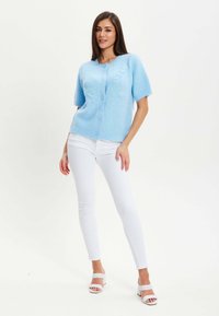 Light blue knitted cardigan with short sleeves, featuring a button closure and textured details. Paired with white pants and heels.