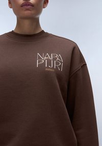 Brun sweatshirt i mjukt tyg, med rund halsringning och tryckt vitt logo med geometriska element på framsidan.
