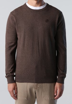 Mężczyzna w brązowym swetrze z długim rękawem na białej podkoszulce i beżowych spodniach, stojący z ramionami swobodnie opuszczonymi wzdłuż ciała.