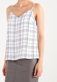 Bílý bezrukávový top s černým mřížkovaným vzorem, V- výstřihem a nastavitelnými tenkými ramínky, kombinovaný se šedou přiléhavou sukni.