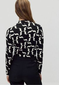 Långärmad skjorta med ett vågigt svart och vitt mönster, tillverkad av ett mjukt material. Baksidan har en liten knapppdetalj och en rundad halsringning.