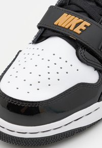 Zbliżenie czarno-białego buta Nike z perforowaną białą skórą na czubku oraz czarnym paskiem z pomarańczowym logo Nike.