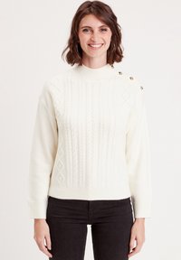 Pull en tricot couleur crème avec un col roulé côtelé, des motifs en câble texturés et des boutons métalliques sur l'épaule gauche.