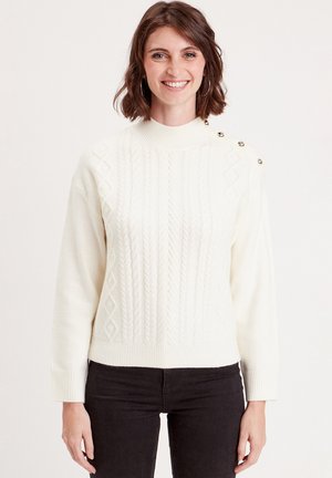 Pull en tricot couleur crème avec un col roulé côtelé, des motifs en câble texturés et des boutons métalliques sur l'épaule gauche.