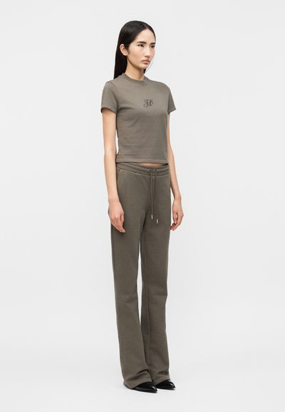 Νέα γυναίκα wearing crop top με κοντά μανίκια σε ελαιόχρωμο και φαρδιά φούτερ παντελόνια στο ίδιο χρώμα, στέκεται πλαγίως μπροστά σε ένα μονόχρωμο φόντο.