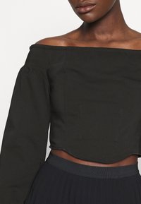 Top corto nero off-the-shoulder con maniche lunghe puffate e due tasche frontali; presenta un design strutturato e una texture del tessuto liscia.