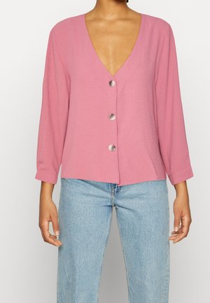 Frau trägt einen rosa langärmeligen Cardigan mit drei Knöpfen und hellblaue hoch taillierte Jeans vor einfarbigem Hintergrund.