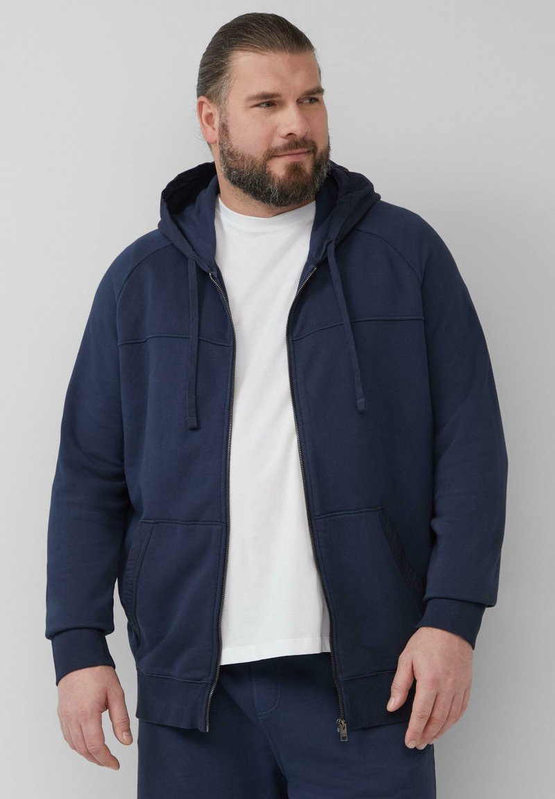 Marineblaues Zip-Hoodie aus einem weichen Baumwollmix, ausgestattet mit einer kleinen Kapuze, Fronttaschen und gerippten Bündchen, getragen über einem weißen T-Shirt.