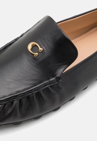 Sorte læderslippers med et dekorativt guldemblem, en blød tekstureret overdel og et struktureret design med en afrundet tå.