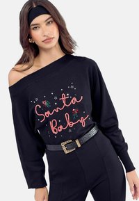 Fekete off-shoulder pulóver, amelyen piros zsinórral van felírva a "Santa Baby" felirat, virágos díszítéssel és ezüst csillag díszekkel, fekete övvel párosítva.