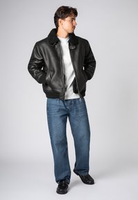 Man staat met zijn handen in zijn zakken, draagt een zwarte leren jas met een bontkraag, een wit shirt, blauwe jeans en zwarte schoenen, en kijkt opzij.