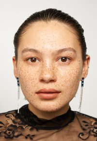 Ansiktsportrett som viser en modell med fremtredende fregner, iført en svart blonder topp og lange sølvøredobber med blå detaljer.