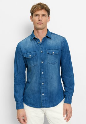Mann trägt ein mittelblaues Jeanshemd mit Brusttaschen und weiße Hose, steht vor einem schlichten weißen Hintergrund.