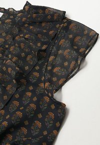 Blouse noire transparente avec un motif floral orange et vert, ornée de volants le long du décolleté et de manches courtes légèrement évasées.