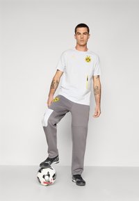 Lysgrå kortærmet skjorte med gule detaljer og logo, parret med grå sweatpants med et hvidt panel og sidelommer, stående på en fodbold.
