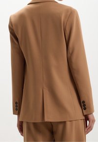 Blazer marron sur mesure avec une fente unique à l'arrière, des revers crantés, et quatre boutons noirs décoratifs sur les manches. Texture de tissu lisse.