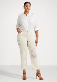 Jeans beige de cintura alta con estampados decorativos en púrpura y azul. Combinados con una camisa blanca de lino con botones. Tacones marrones con tiras.