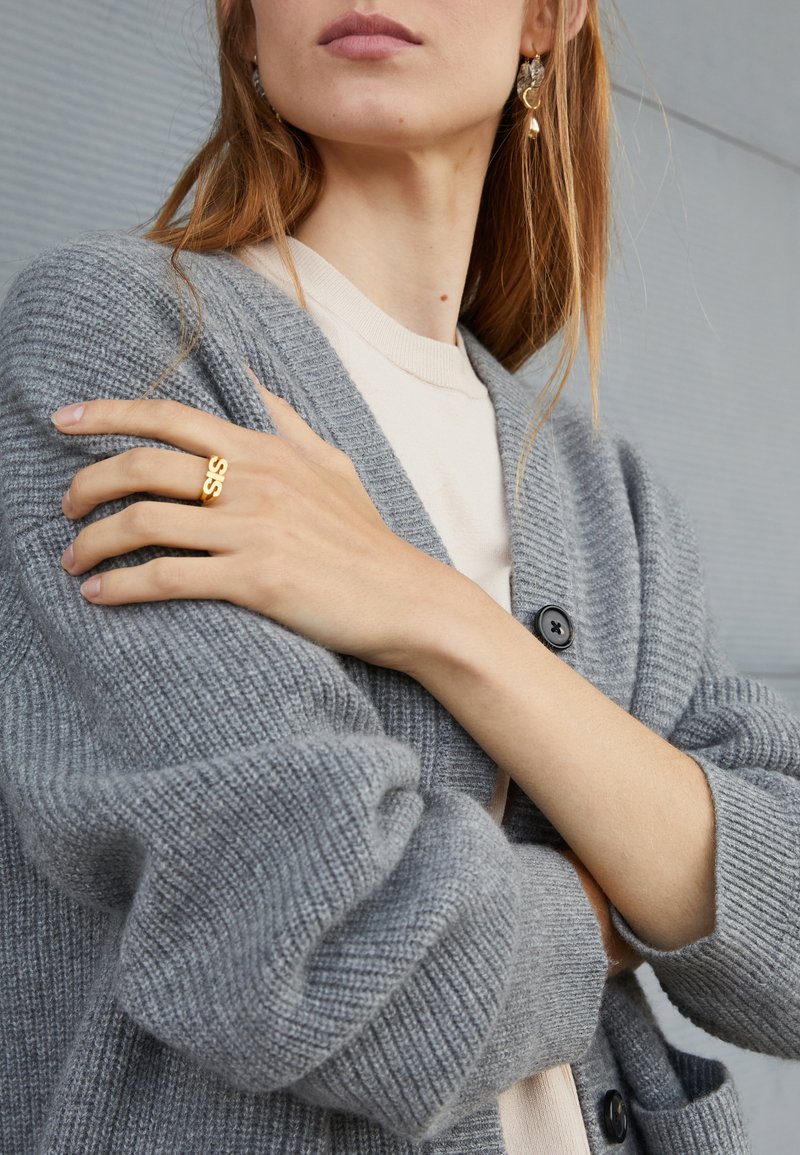 Világos bőrű nő, bézs felsőt és szürke kötött kardigánt visel, a bal kezén arany "SIS" gyűrűt, a bal fülén pedig szív alakú fülbevalót mutat.