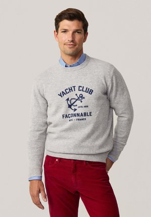 Suéter gris de punto con un estampado gráfico en azul marino de "Yacht Club Faconnable" y un diseño de ancla, combinado con pantalones de pana rojos.