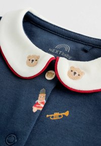 Navy Baby-Shirt mit einem weißen Kragen, der rot eingefasst ist, und mit gestickten Bären sowie einem Spielzeugsoldaten mit Trompeten-Motiv.