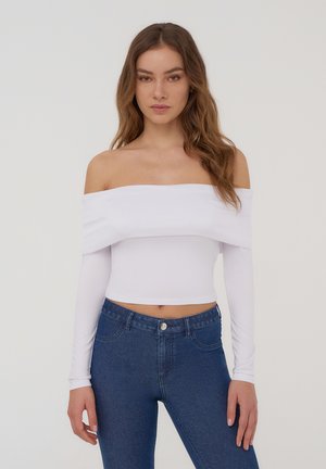 Top corto bianco a spalle scoperte con maniche lunghe, realizzato in tessuto elasticizzato, abbinato a jeans blu a vita alta. Texture semplice, aspetto morbido.