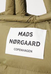 Borsa imbottita verde oliva con due manici superiori, caratterizzata da un'etichetta rettangolare bianca con testo nero: "MADS NØRGAAARD COPENHAGEN."