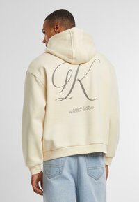 Creme hoodie med et stort gråt "K" logo og teksten "KARMA CLUB" på ryggen. Har en hætte med snoretræk og ribbede manchetter.