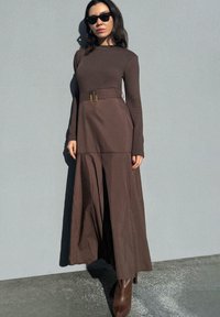 Robe maxi marron à manches longues avec une taille ceinturée, fabriquée en tissu lisse ; présente une jupe fluide et des détails de couture subtils. Associée à des bottes marron.