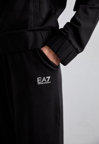 Crne atletske hlače od mekog materijala, s bočnim džepom i bijelim EA7 Emporio Armani logotipom na lijevom bedru.