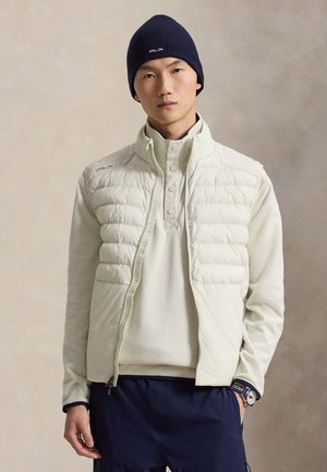 Homme portant un bonnet bleu marine, un gilet matelassé blanc sur un pull blanc, un pantalon bleu marine et une montre avec un bracelet visible.