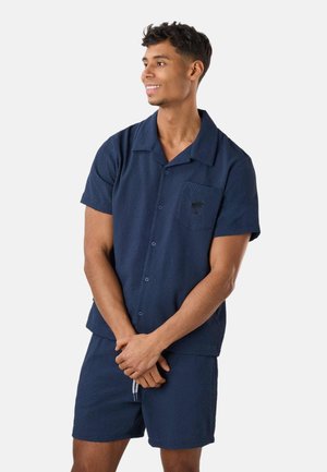 Jeune homme souriant, portant une chemise texturée bleu marine à manches courtes boutonnée et un short assorti, debout les mains jointes devant.
