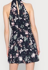 Blumenkleid mit Halsband in Dunkelblau, das weiße und rosa Blumenmuster, einen Schlüssellochausschnitt hinten und eine taillierte Taille mit einem schwingenden Rock zeigt.