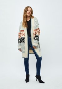 Cardigan en maille de couleur crème avec des motifs géométriques en bleu, noir et orange. Il présente un devant ouvert et des manches longues. Porté par-dessus un haut noir.