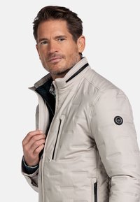 Lichtbeige jas met een gestructureerd patroon, hoge kraag, ritssluiting aan de voorkant en accenten bij de zakken. Voorzien van een zwart logopatch op de mouw.