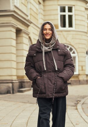 Une femme souriante se tient sur un trottoir en ville, vêtue d’un manteau matelassé foncé et d’une écharpe à capuche grise.