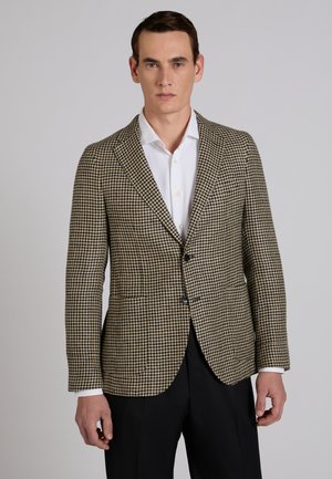 Mężczyzna w beżowo-czarnej marynarce w jodełkę, na białej koszuli i czarnych spodniach, stojący na tle jednolitego szarego tła.