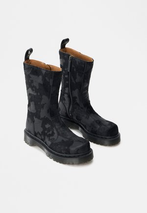 Par de botas pretas até meio da canela com padrão texturizado, fechos laterais, solas grossas e alças para puxar, sobre fundo branco.