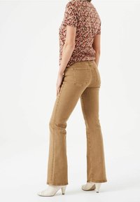 Zahnuté béžové džíny s jemnou strukturou, s kapsami na zadní straně. Modelka má na sobě vzorovaný top a bílé podpatky, pohled zezadu.