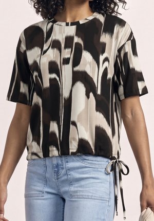 Vrouw die een abstract zwart, wit en bruin patroon kortemouwtop met een zijstrikdetail en lichtblauwe hoogwaterjeans draagt.