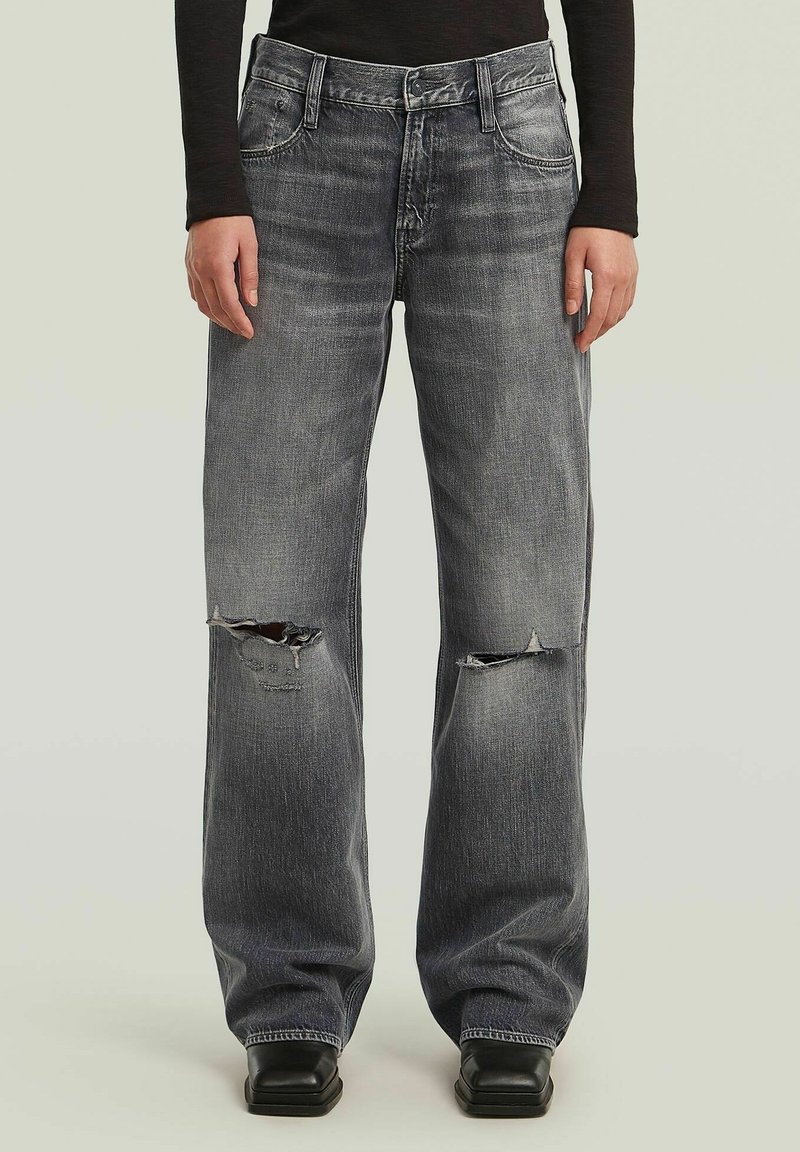 Personne portant un jean noir délavé et ample avec des déchirures aux deux genoux, associé à des chaussures noires à bout carré et un haut noir à manches longues.