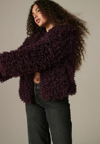 Lilla faux fur jakke med en firkantet skæring, der har en tekstureret, krøllet overflade og brede ærmer. Kombinationen med højtaljede mørke jeans.